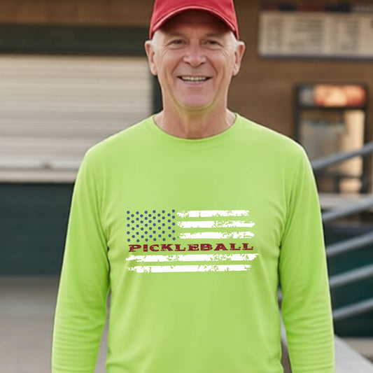 A man wearing a lime green short-sleeve t-shirt with a pickleball graphic of a distressed flag made out of pickleballs and the text 'PICKLEBALL' replacing one of the stripes, standing on a pickleball court.