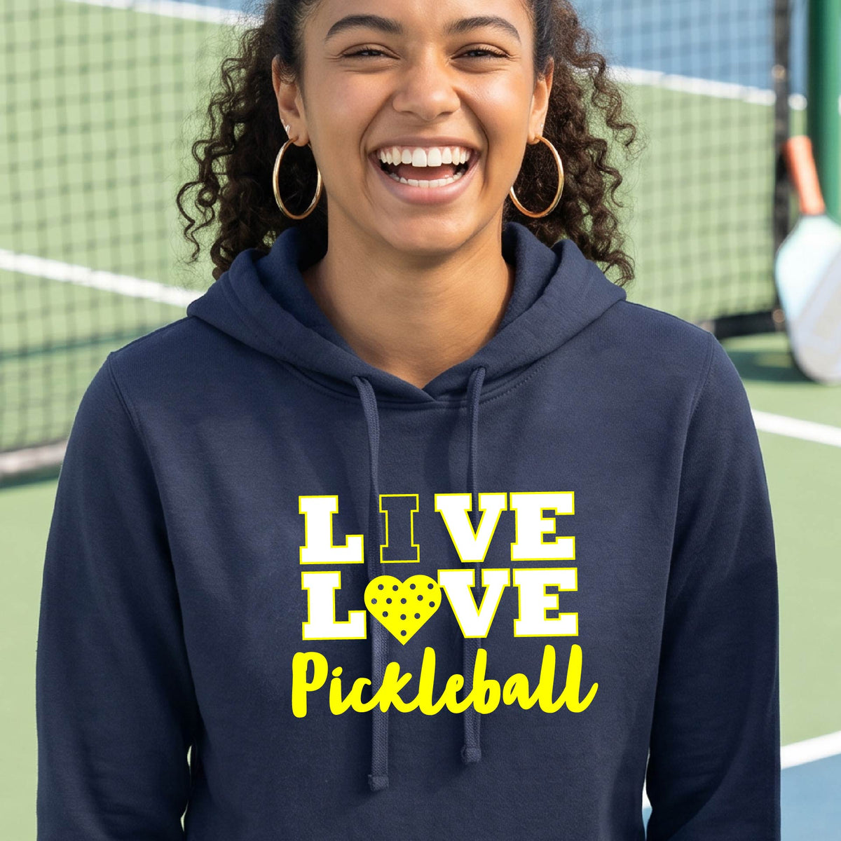 A smiling blonde woman wearing a navy blue pullover hoodie with a yellow graphic that reads 'I LOVE PICKLEBALL' with a heart-shaped pickleball motif, photographed on a pickleball court with a chain-link fence in the background.