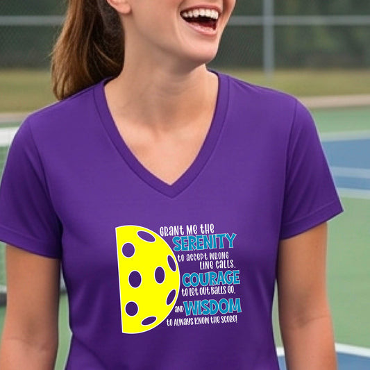 A woman on a pickleball court wearing a purple short-sleeve v-neck t-shirt with a graphic of the Serenity Prayer made for pickleball: “Grant me the serenity to Accept Wrong Line Calls; Courage to Let Out Balls Go; and, Wisdom to Always Know the Score” next to a half-pickleball icon, standing on a pickleball court.