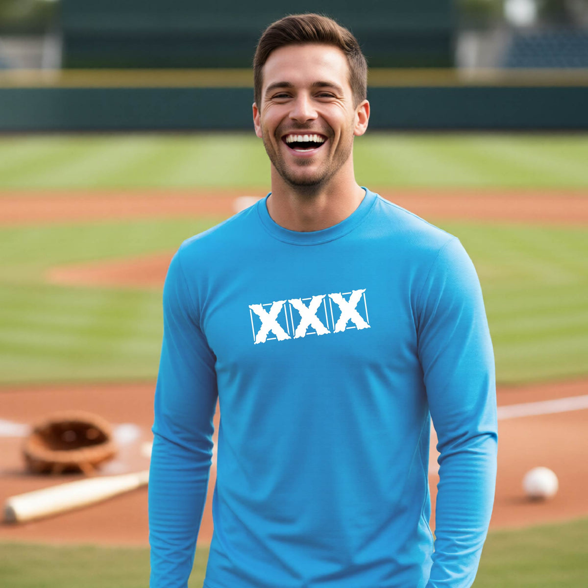 A smiling man wearing a bright blue long-sleeve athletic shirt with a white XXX graphic on the chest, standing on a baseball field.