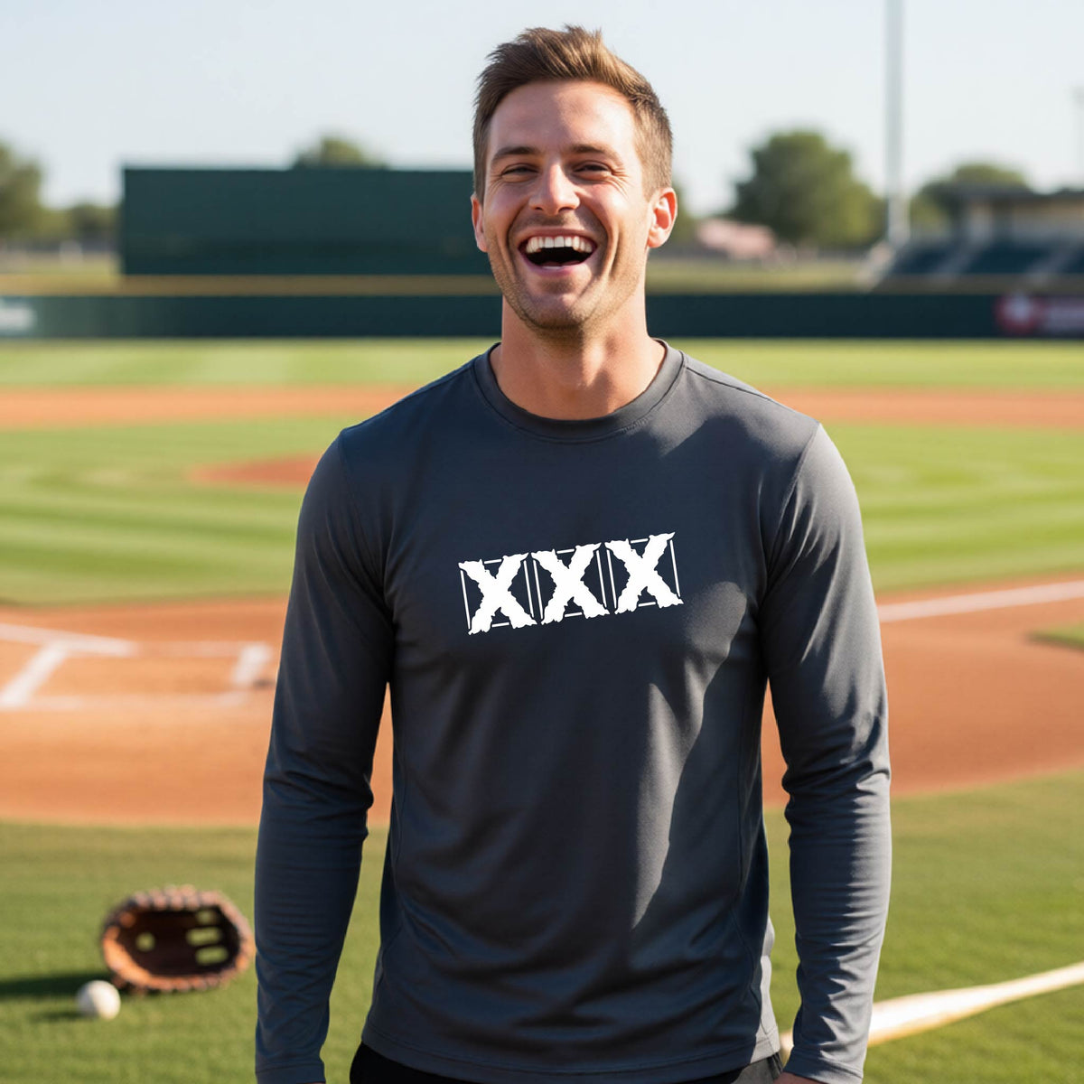 A man laughing on a baseball field wearing a dark gray long-sleeve shirt with a white XXX graphic across the chest.