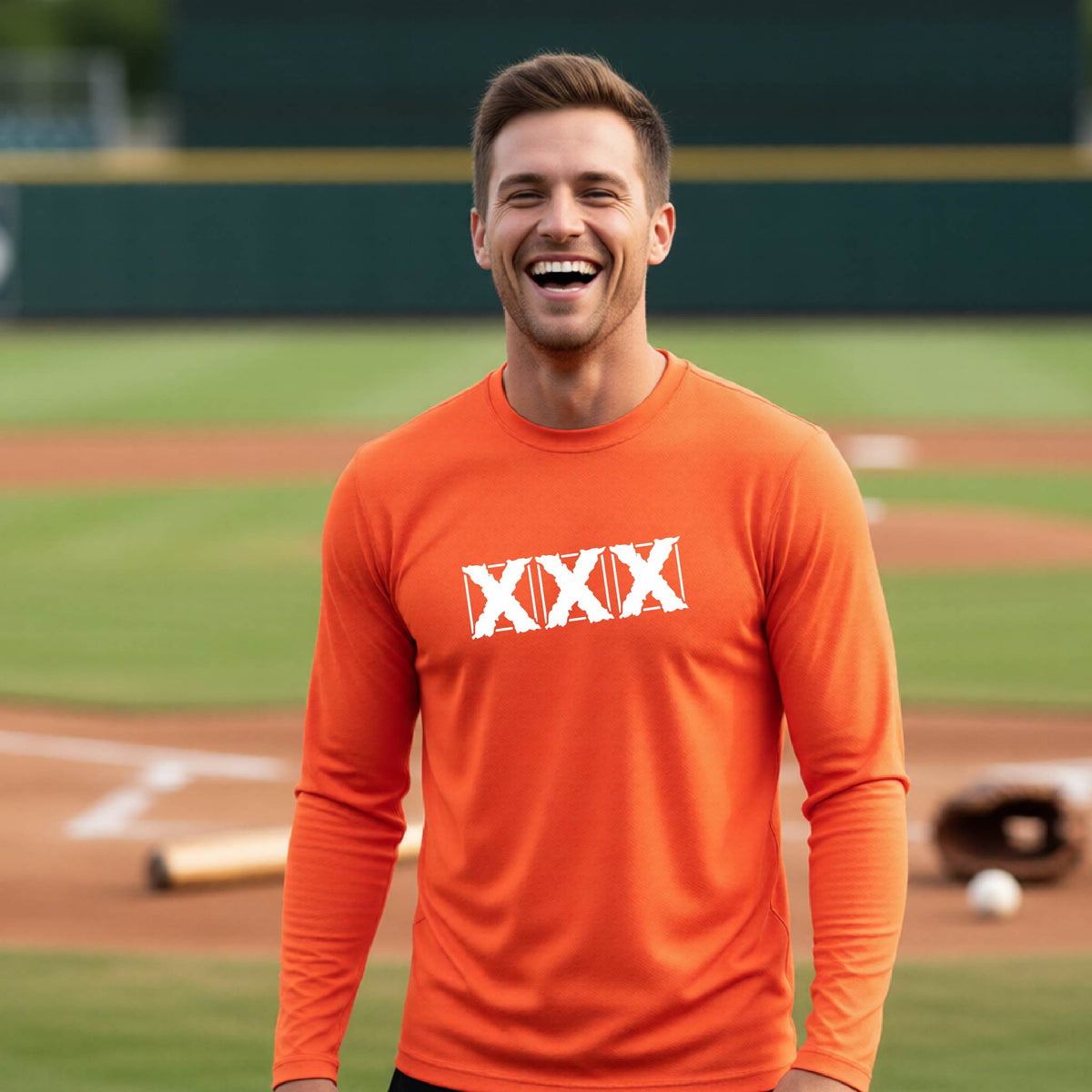 A man smiling on a baseball field wearing a bright orange long-sleeve athletic shirt with a white XXX logo printed across the chest.