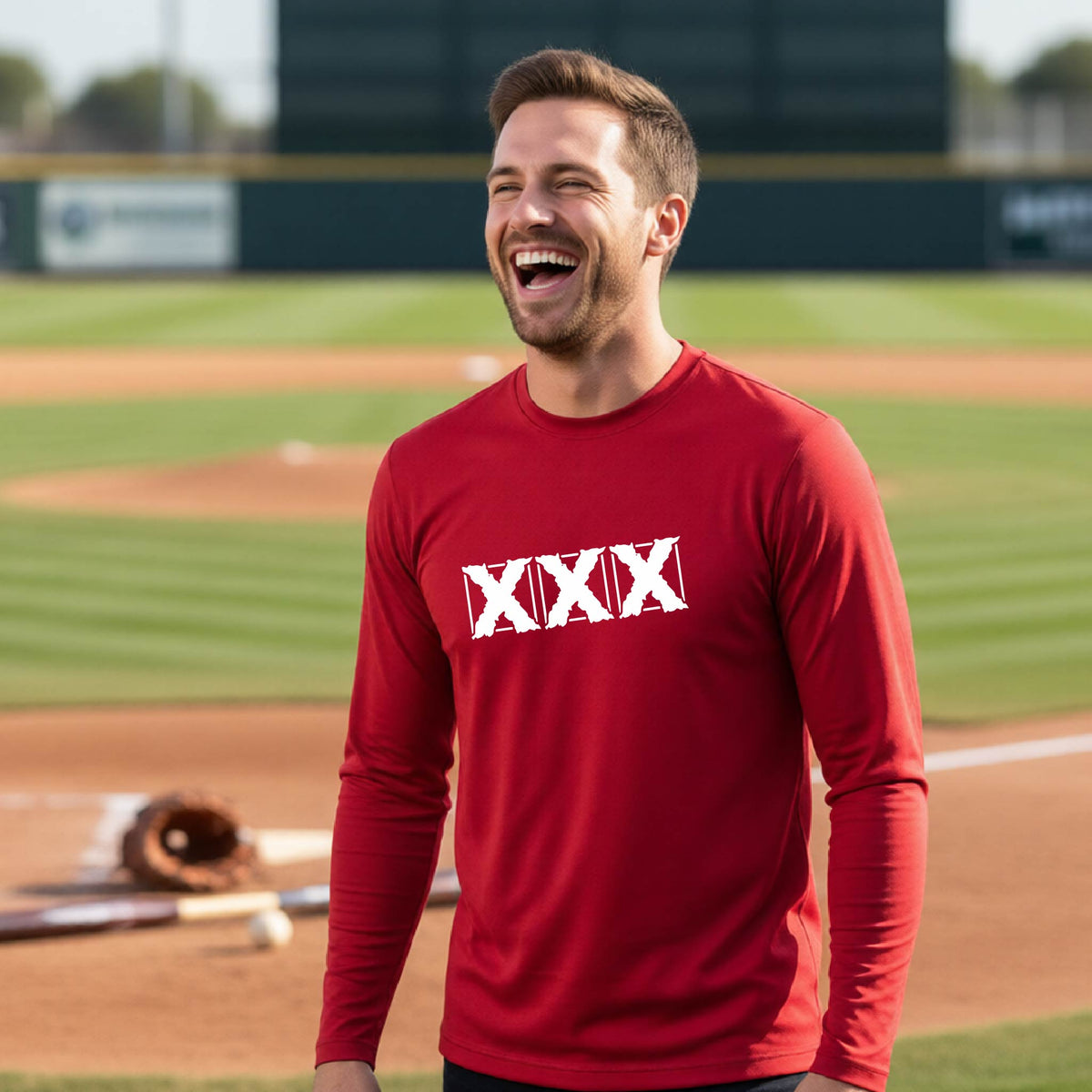 A man laughing on a baseball field wearing a red long-sleeve shirt with a white XXX logo across the chest.