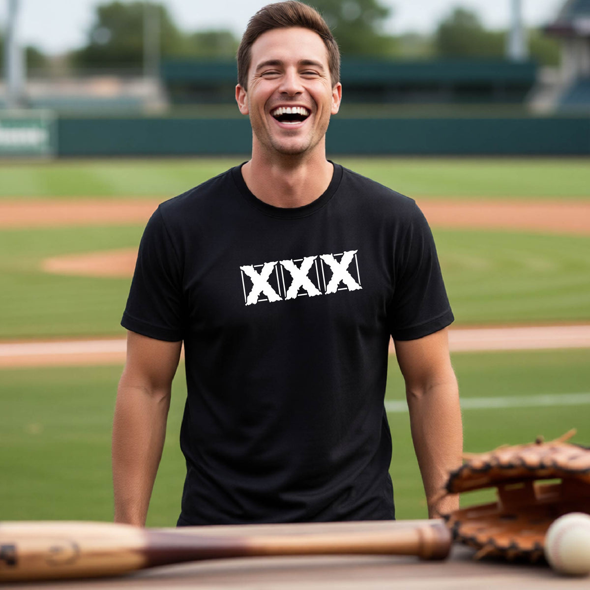 A man laughing on a baseball field wearing a black t-shirt with a white XXX graphic across the chest, with a baseball bat, glove, and ball in the foreground.