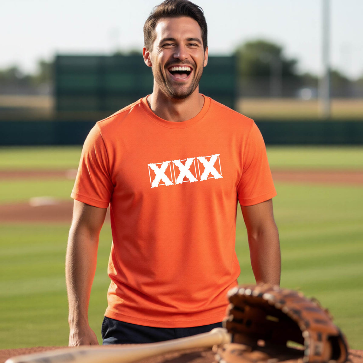 A man laughing on a baseball field wearing an orange t-shirt with a white 'XXX' baseball graphic on the chest, with a baseball field in the foreground.