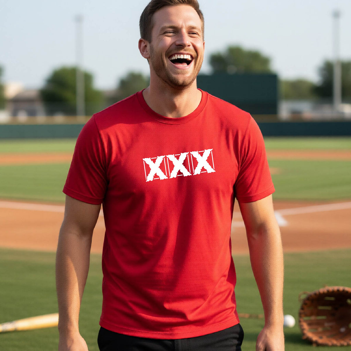 A man laughing on a baseball field wearing a red t-shirt with a white 'XXX' graphic across the chest.