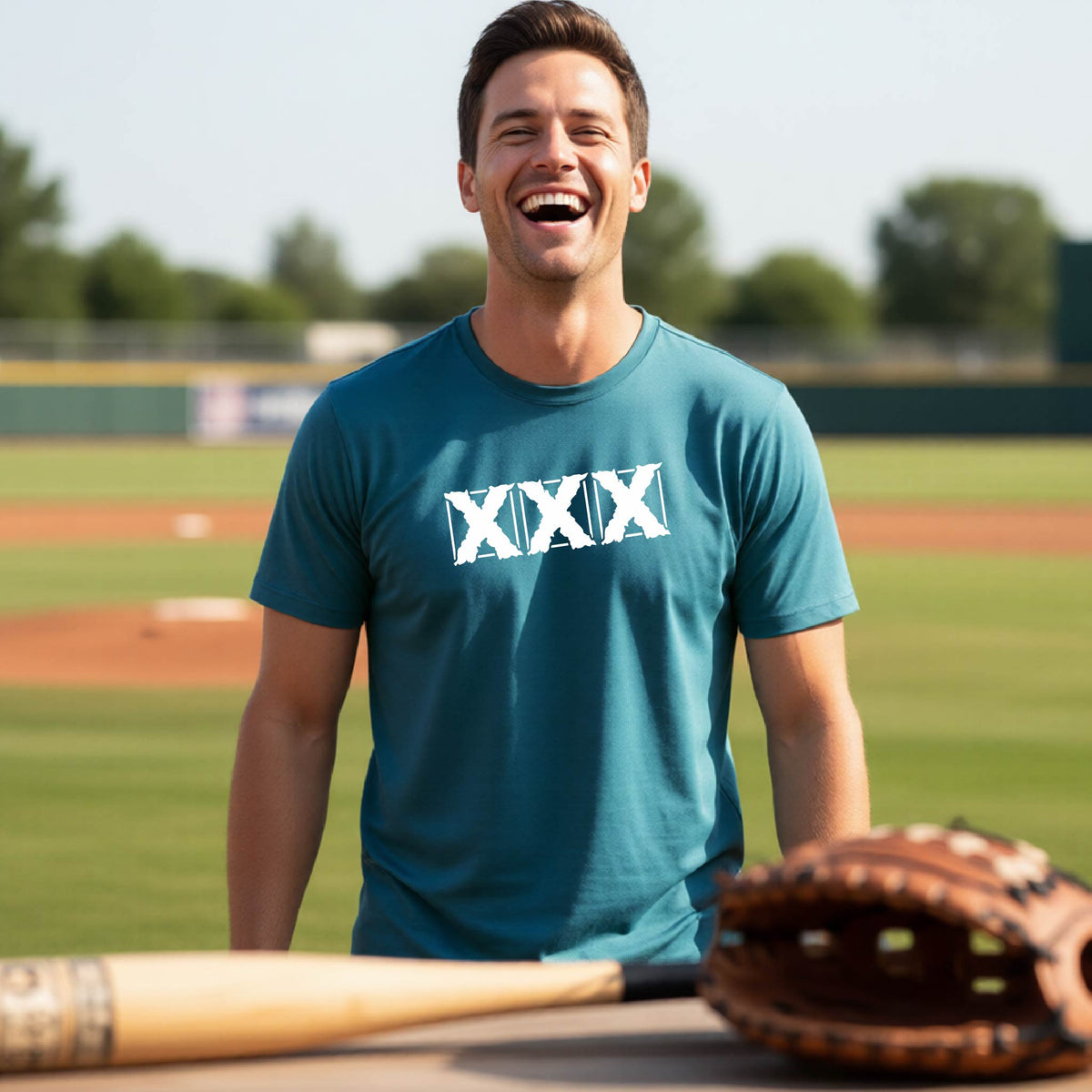 A man laughing on a baseball field wearing a teal t-shirt with a white XXX graphic across the chest, with a baseball bat and glove in the foreground.