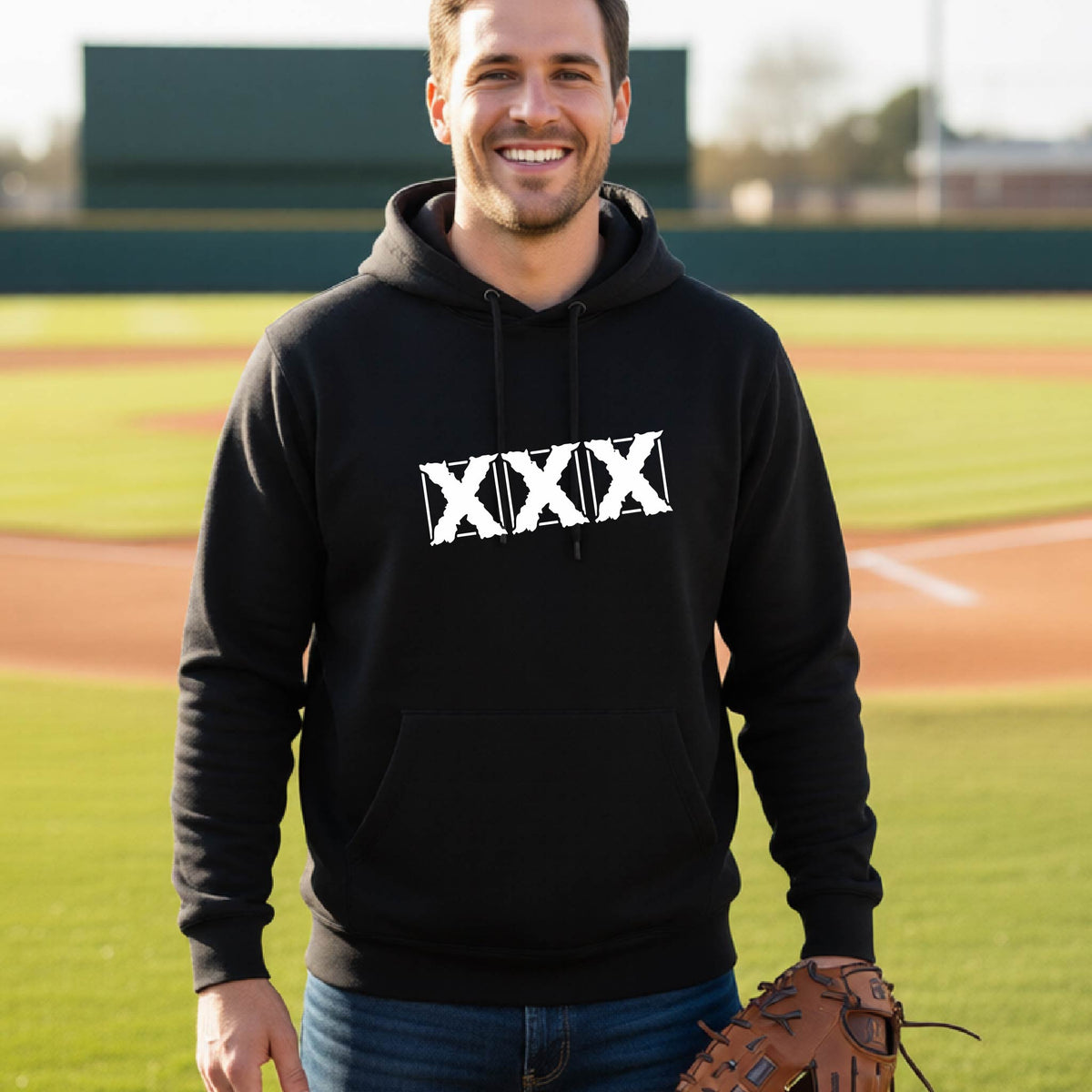 A man wearing a black hoodie with a white distressed 'XXX' graphic on the chest, standing on a baseball field and holding a brown leather glove.