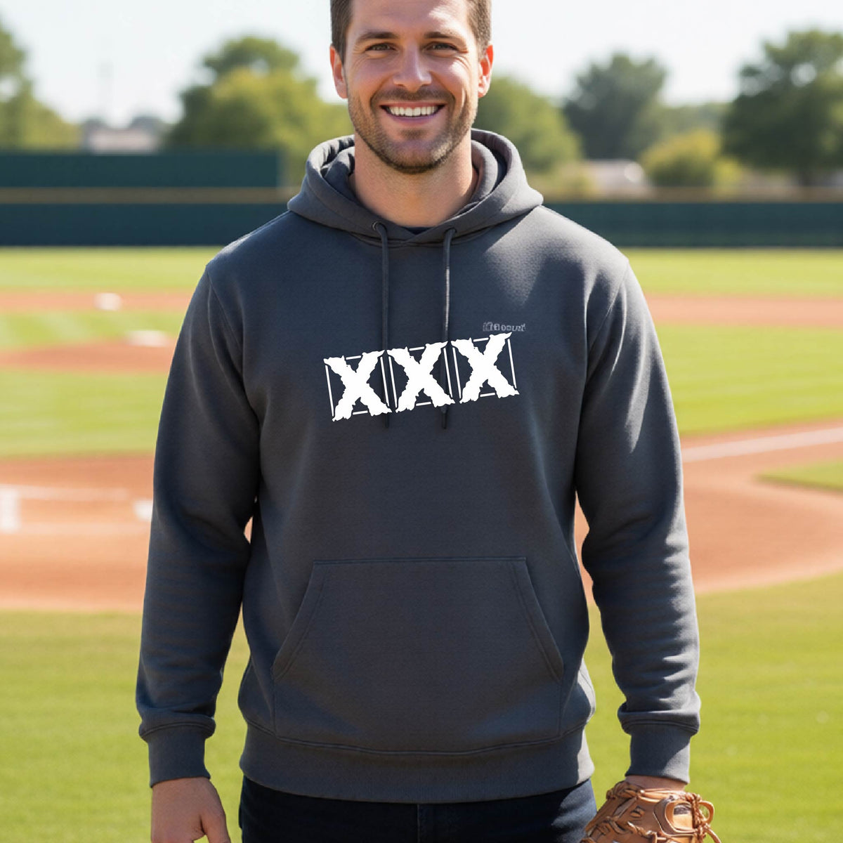 A man wearing a dark gray hoodie with a white distressed 'XXX' graphic on the chest stands on a baseball field, holding a brown baseball glove.