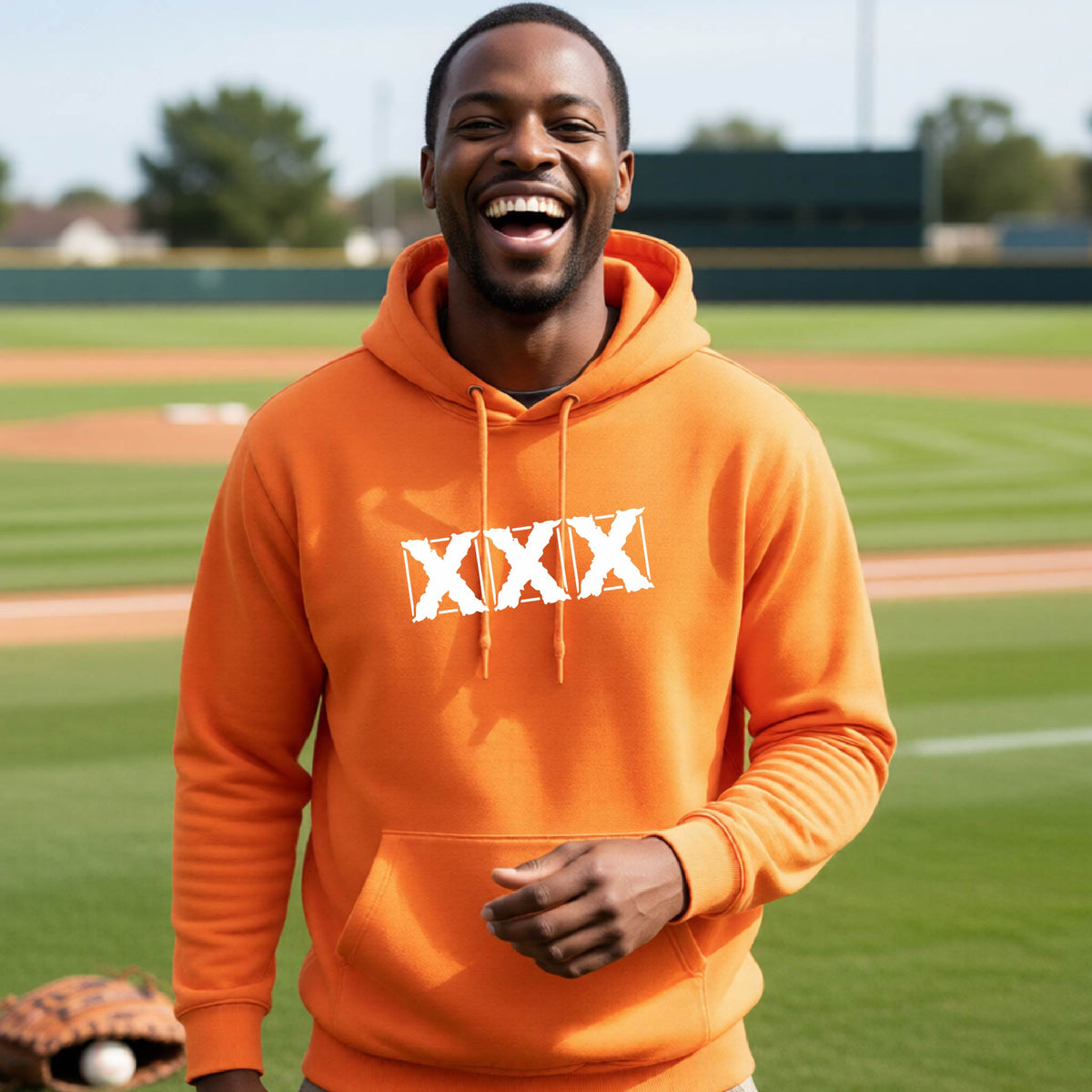 A man wearing a bright orange hoodie with a white 'XXX' graphic on the chest, standing on a baseball field.