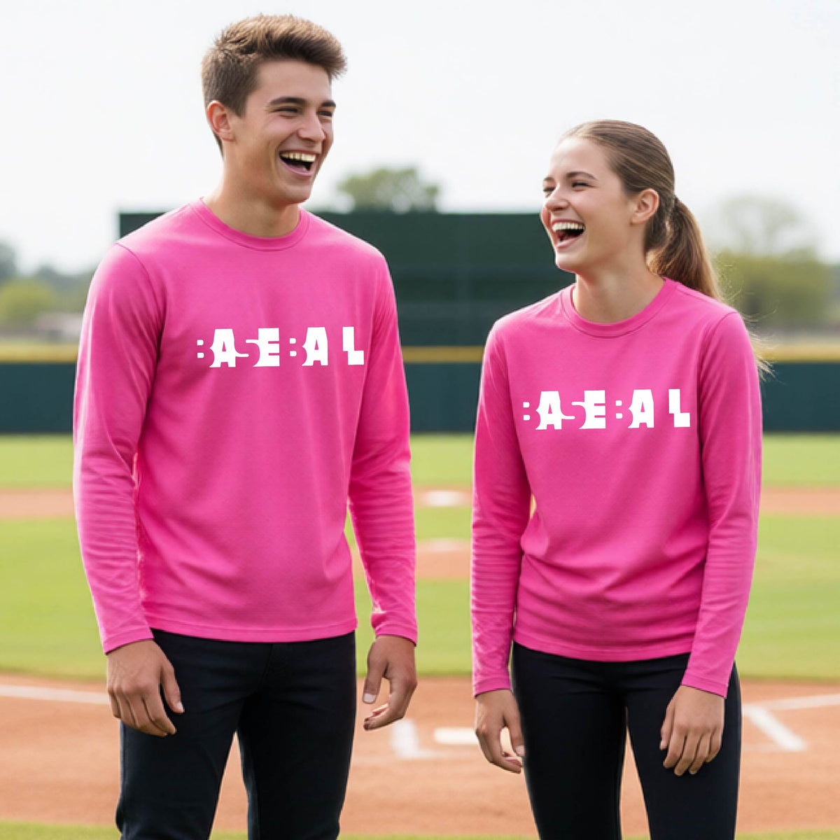 Two young athletes, standing on a baseball field wearing matching bright pink long-sleeve t-shirts with white pixelated text that reads 'BASEBALL' in a cutout letter font.