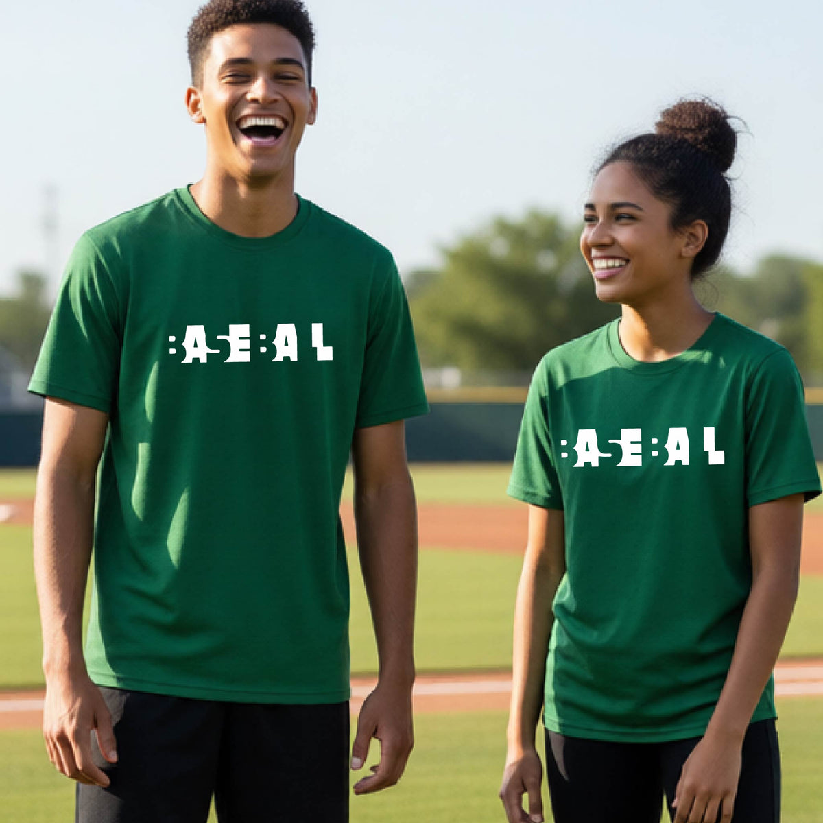 Two young people standing on a baseball field wearing matching green t-shirts with the text 'BASEBALL' in a cutout artsy design printed in white across the chest.