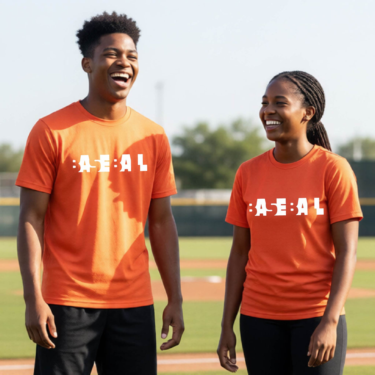 Two young people standing on a baseball field wearing matching orange t-shirts with the text 'BASEBALL' in a cutout artsy design printed in white across the chest.