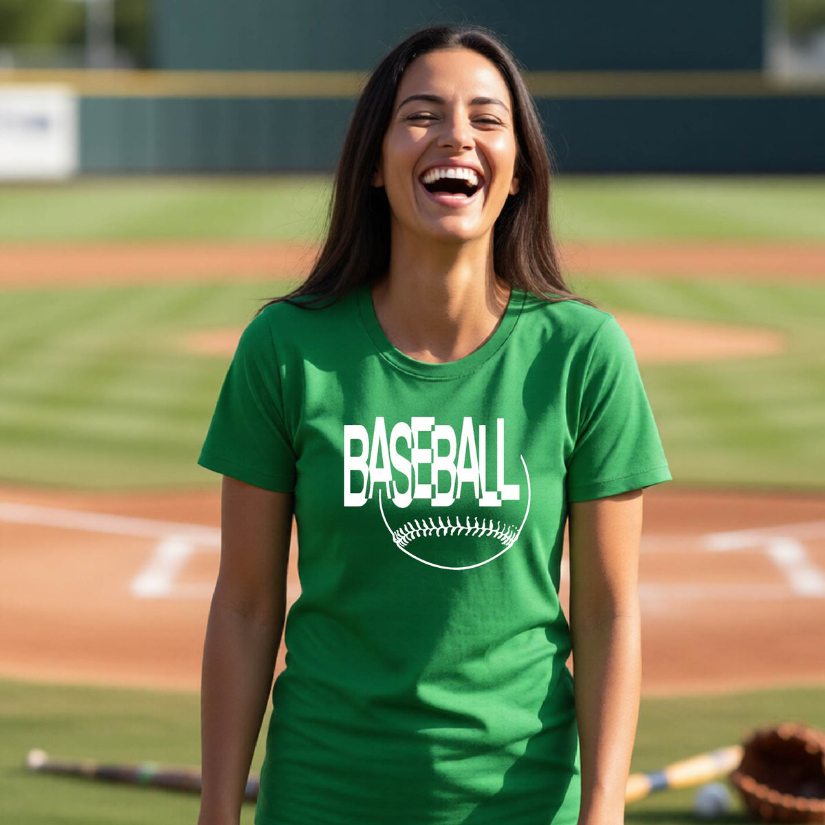 A woman laughing on a baseball field wearing a green short-sleeve t-shirt with a white 'BASEBALL' graphic and baseball stitching design on the front, standing on a baseball field.