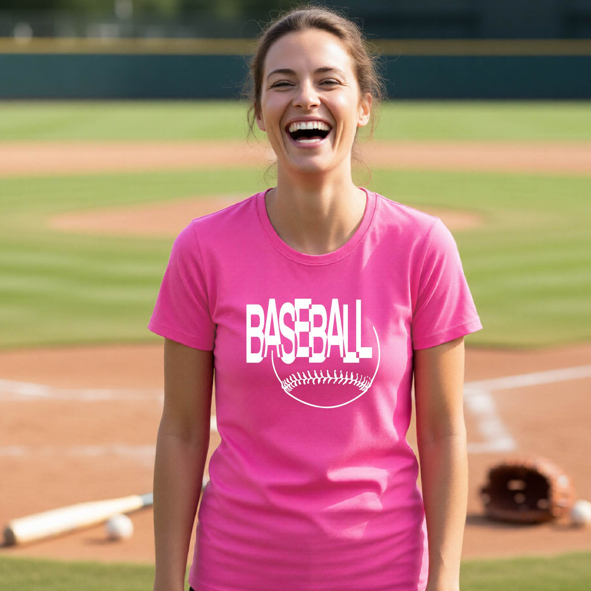 A woman laughing on a baseball field wearing a pink short-sleeve t-shirt with a white 'BASEBALL' graphic and baseball stitching design on the front, standing on a baseball field.