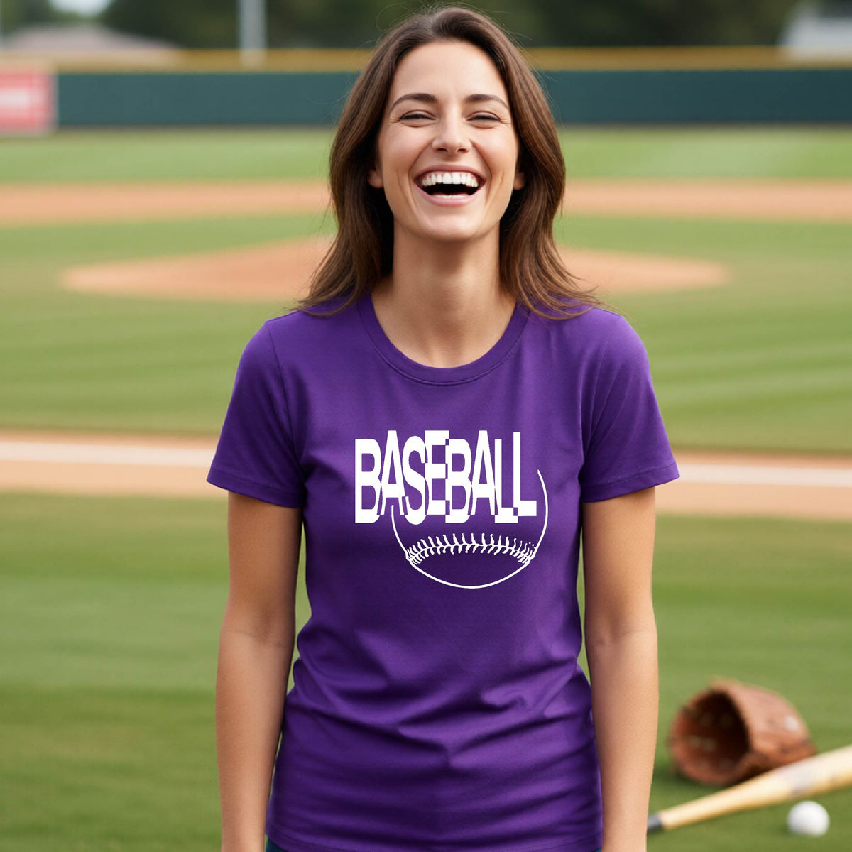 A woman laughing on a baseball field wearing a purple short-sleeve t-shirt with a white 'BASEBALL' graphic and baseball stitching design on the front, standing on a baseball field.