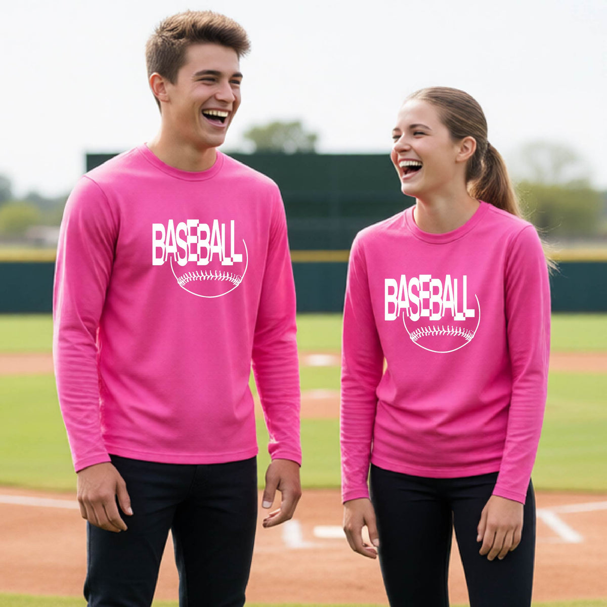 Two young athletes wearing matching pink long-sleeve t-shirts with a white baseball graphic on the chest stands on a baseball field. The shirt features the word 'BASEBALL' in a baseball-style font with a stitched ball design underneath.