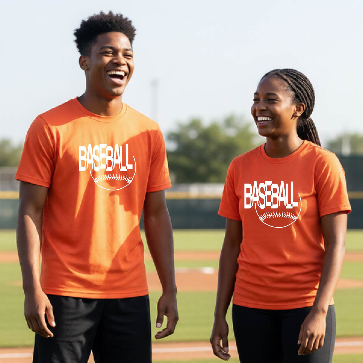 Two young athletes wearing matching orange short-sleeve t-shirts with a white baseball graphic on the chest stands on a baseball field. The shirt features the word 'BASEBALL' in a baseball-style font with a stitched ball design underneath.