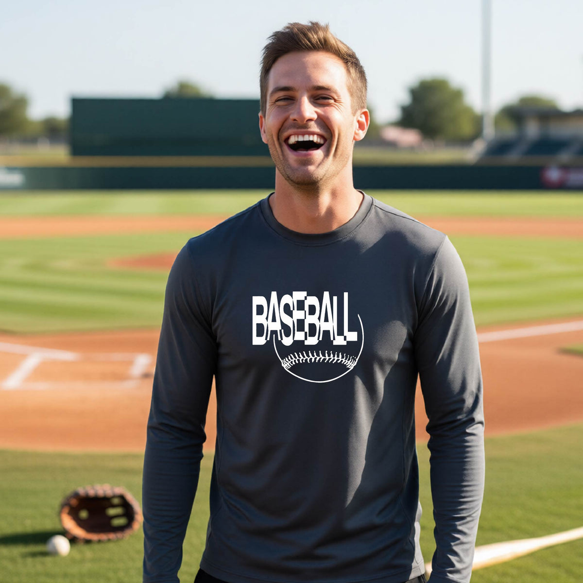 A man laughing on a baseball field wearing a dark gray long-sleeve t-shirt with a white 'BASEBALL' graphic and baseball stitching design on the front, standing on a baseball field.