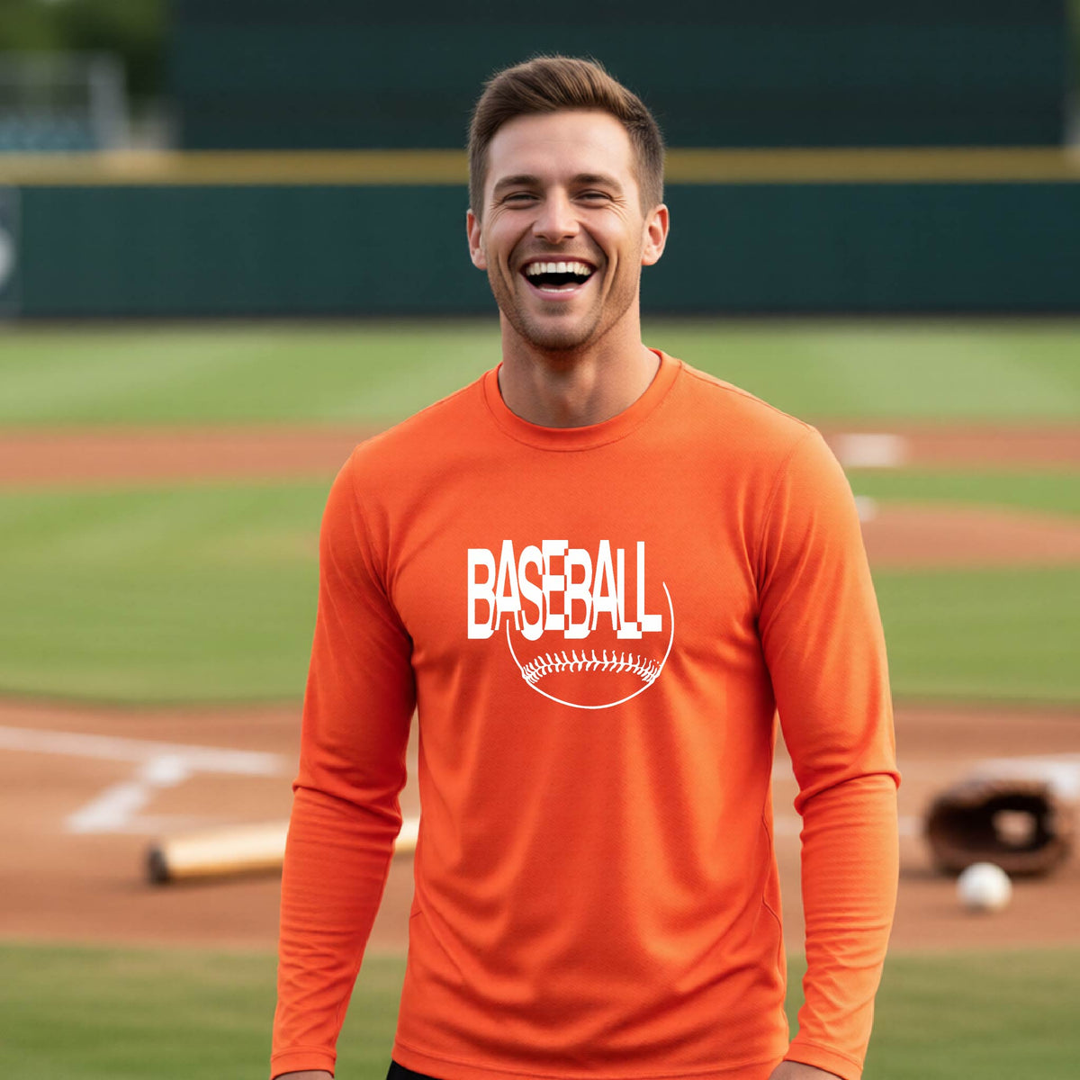 A man laughing on a baseball field wearing an orange long-sleeve t-shirt with a white 'BASEBALL' graphic and baseball stitching design on the front, standing on a baseball field.