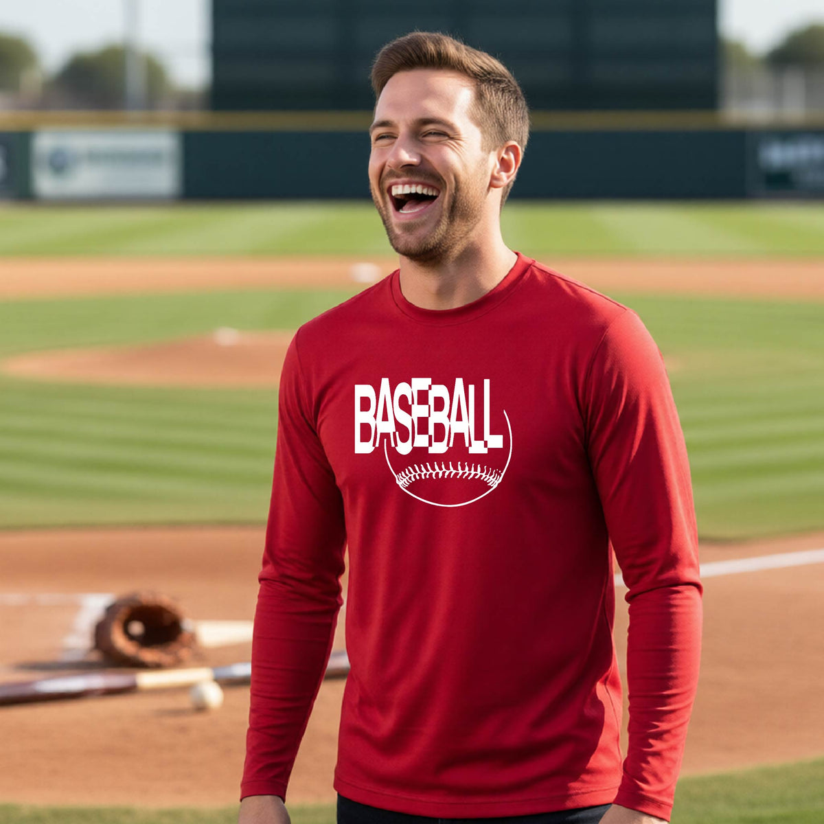 A man laughing on a baseball field wearing a red long-sleeve t-shirt with a white 'BASEBALL' graphic and baseball stitching design on the front, standing on a baseball field.