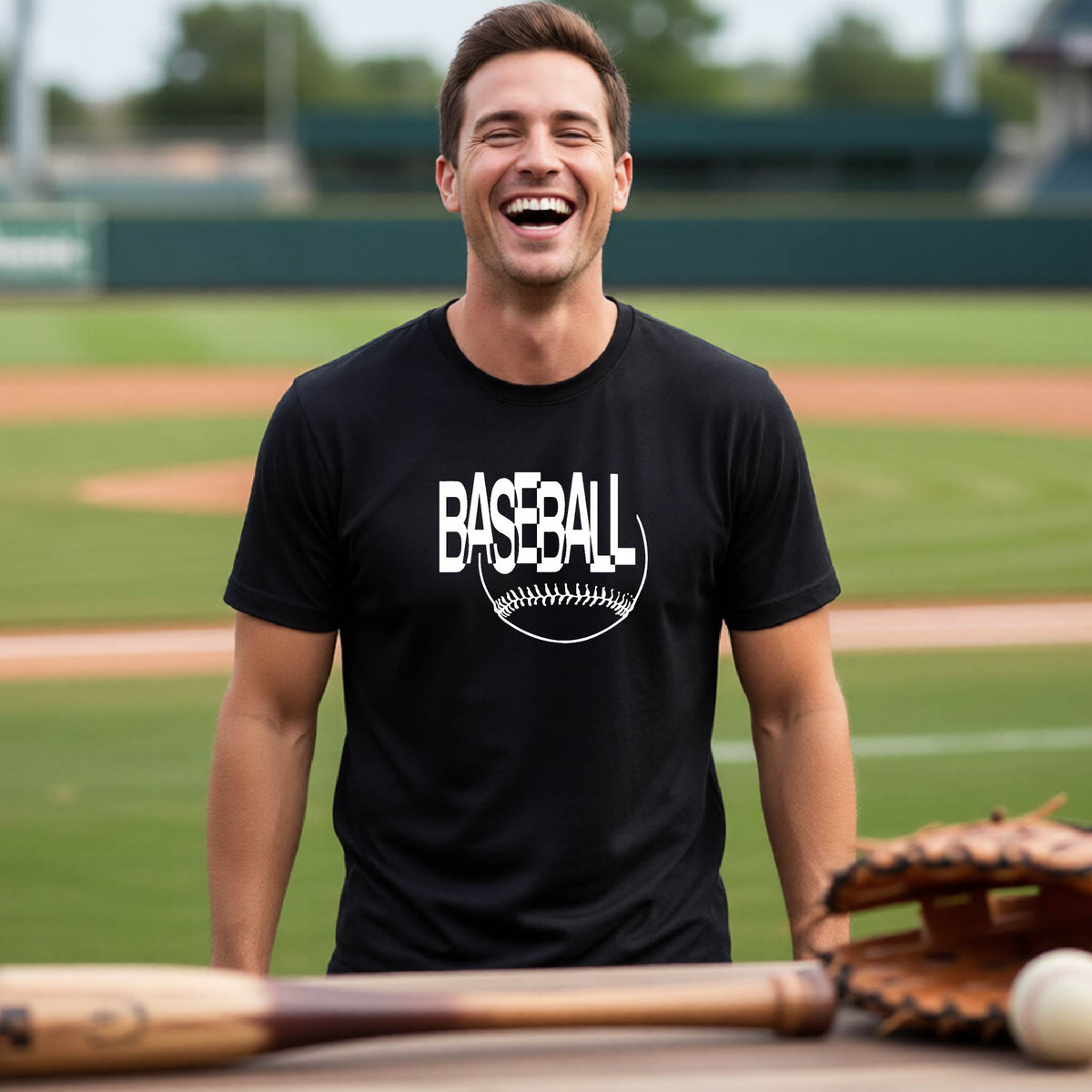 A man laughing on a baseball field wearing a black t-shirt with a white 'BASEBALL' graphic. A baseball bat, glove, and ball are visible in the foreground.