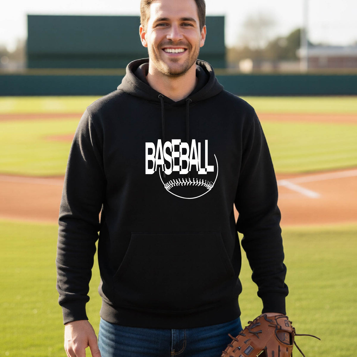 A smiling man wearing a black hoodie with a white baseball graphic on the chest stands on a baseball field. The hoodie features the word 'BASEBALL' in a baseball-style font with a stitched ball design underneath.