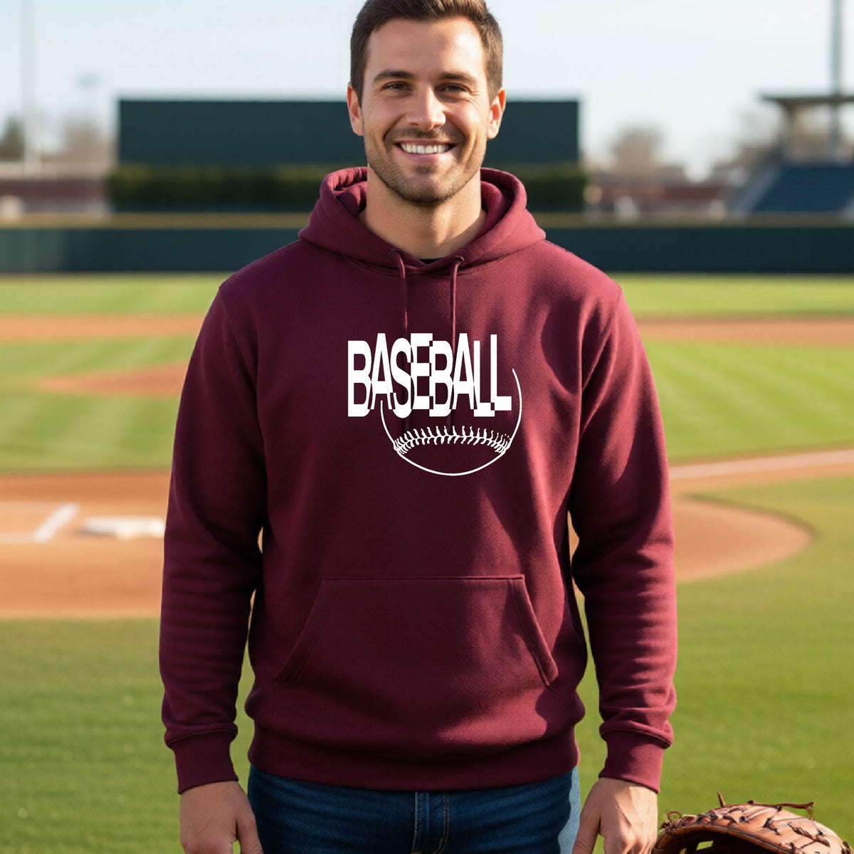 A smiling man wearing a maroon hoodie with a white baseball graphic on the chest stands on a baseball field. The hoodie features the word 'BASEBALL' in a baseball-style font with a stitched ball design underneath.