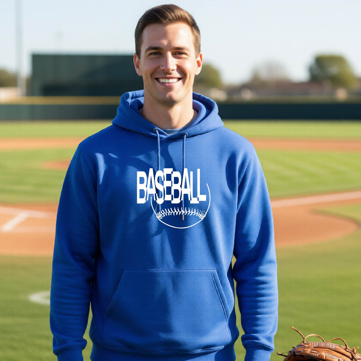 A smiling man wearing a royal blue hoodie with a white baseball graphic on the chest stands on a baseball field. The hoodie features the word 'BASEBALL' in a baseball-style font with a stitched ball design underneath.