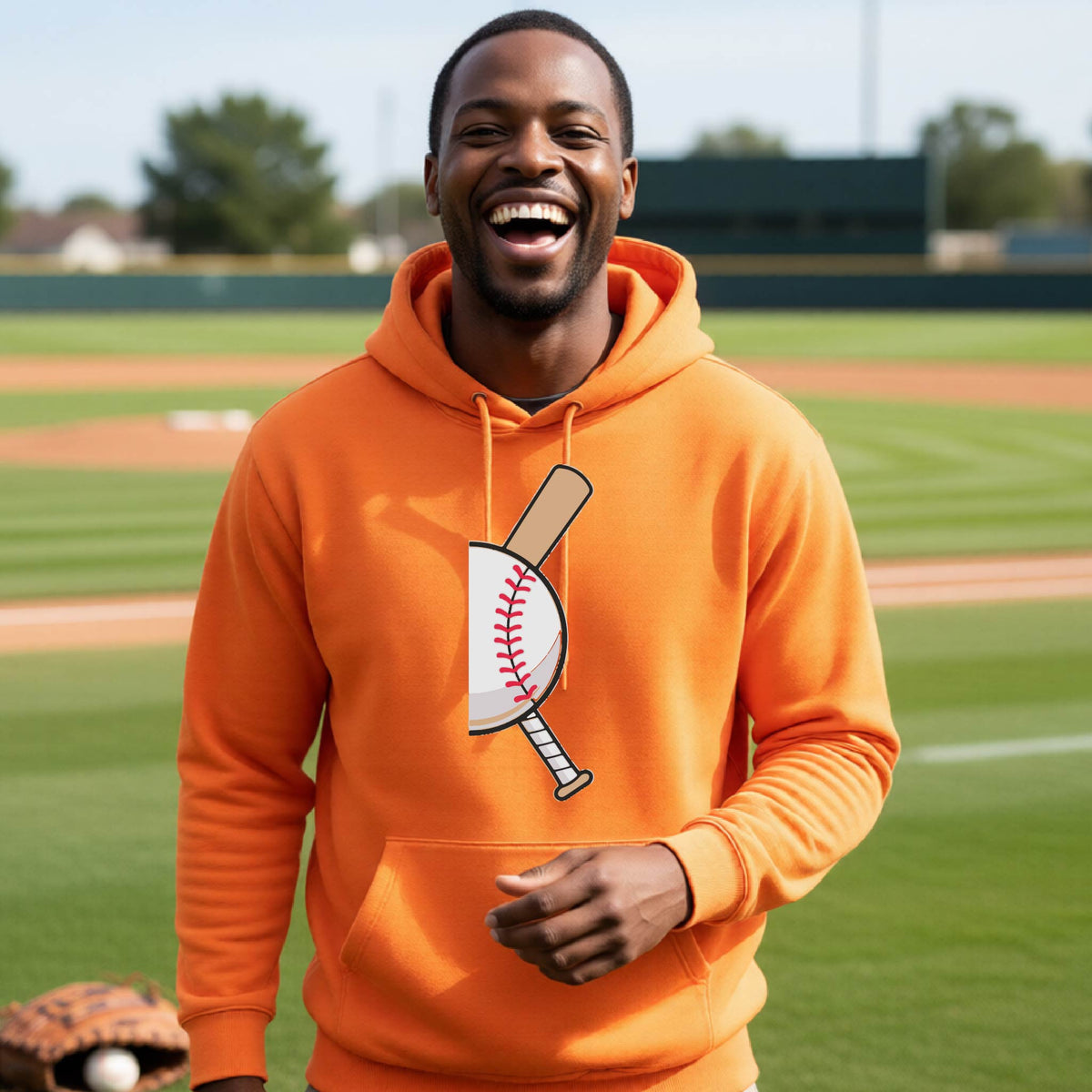 A man smiling in front of a baseball diamond wearing an orange hoodie pullover, with a colorful graphic of a half baseball with a curved bat behind it, printed on the front of the shirt. 