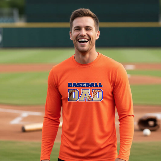 A man smiling in front of a baseball diamond wearing an orange long-sleeve t-shirt with a colored graphic of 'BASEBALL DAD' printed on the front.