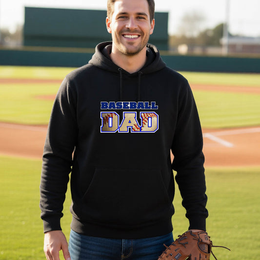 A man smiling in front of a baseball diamond wearing a black hoodie pullover with a colored graphic of 'BASEBALL DAD' printed on the front.