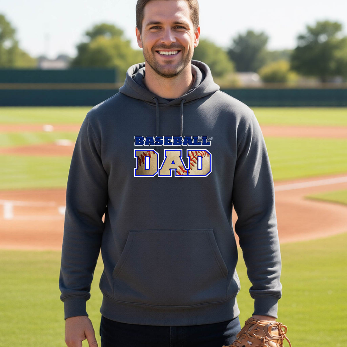A man smiling in front of a baseball diamond wearing a dark gray hoodie pullover with a colored graphic of 'BASEBALL DAD' printed on the front.