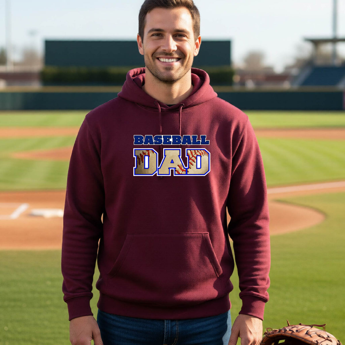 A man smiling in front of a baseball diamond wearing a maroon hoodie pullover with a colored graphic of 'BASEBALL DAD' printed on the front.