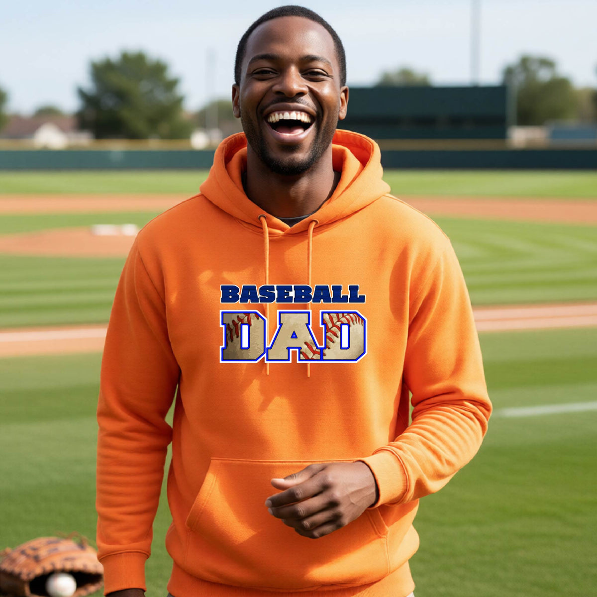 A man smiling in front of a baseball diamond wearing an orange hoodie pullover with a colored graphic of 'BASEBALL DAD' printed on the front.