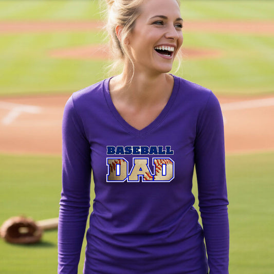 A woman smiling in front of a baseball diamond wearing a purple long-sleeve shirt with a colored graphic of 'BASEBALL DAD' printed on the front. 