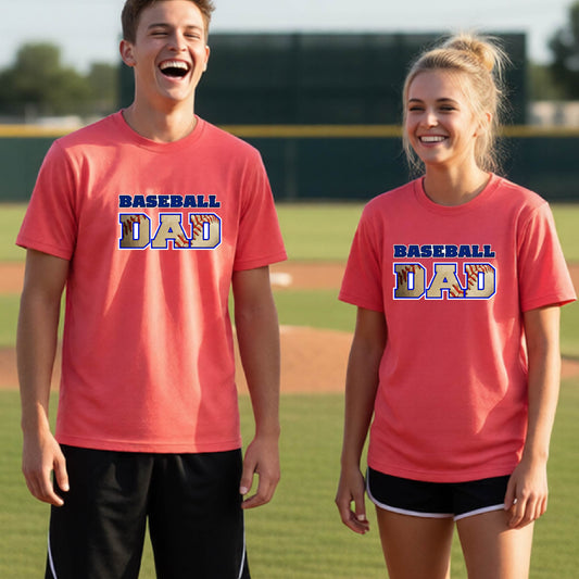 Two young athletes wearing matching coral colored short-sleeve t-shirts smiling in front of a baseball diamond with a colored graphic of 'BASEBALL DAD' printed on the front of the shirts.