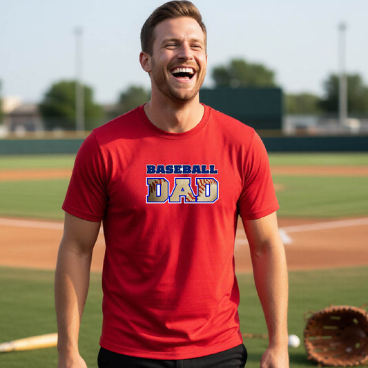 A man smiling in front of a baseball diamond wearing a red short-sleeve t-shirt with a colored graphic of 'BASEBALL DAD' printed on the front.