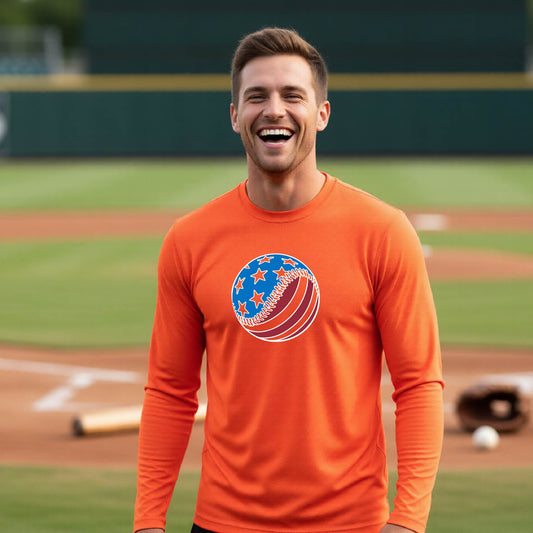 A man smiling in front of a baseball diamond wearing an orange long-sleeve t-shirt with a colored graphic of an American flag patriotic baseball with stars and stripes printed on the front.