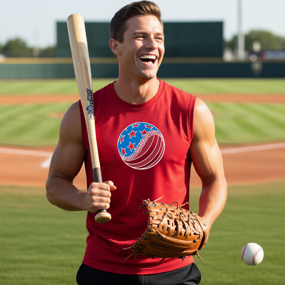 A man smiling in front of a baseball diamond wearing a red sleeveless t-shirt with a colored graphic of an American flag patriotic baseball with stars and stripes printed on the front.