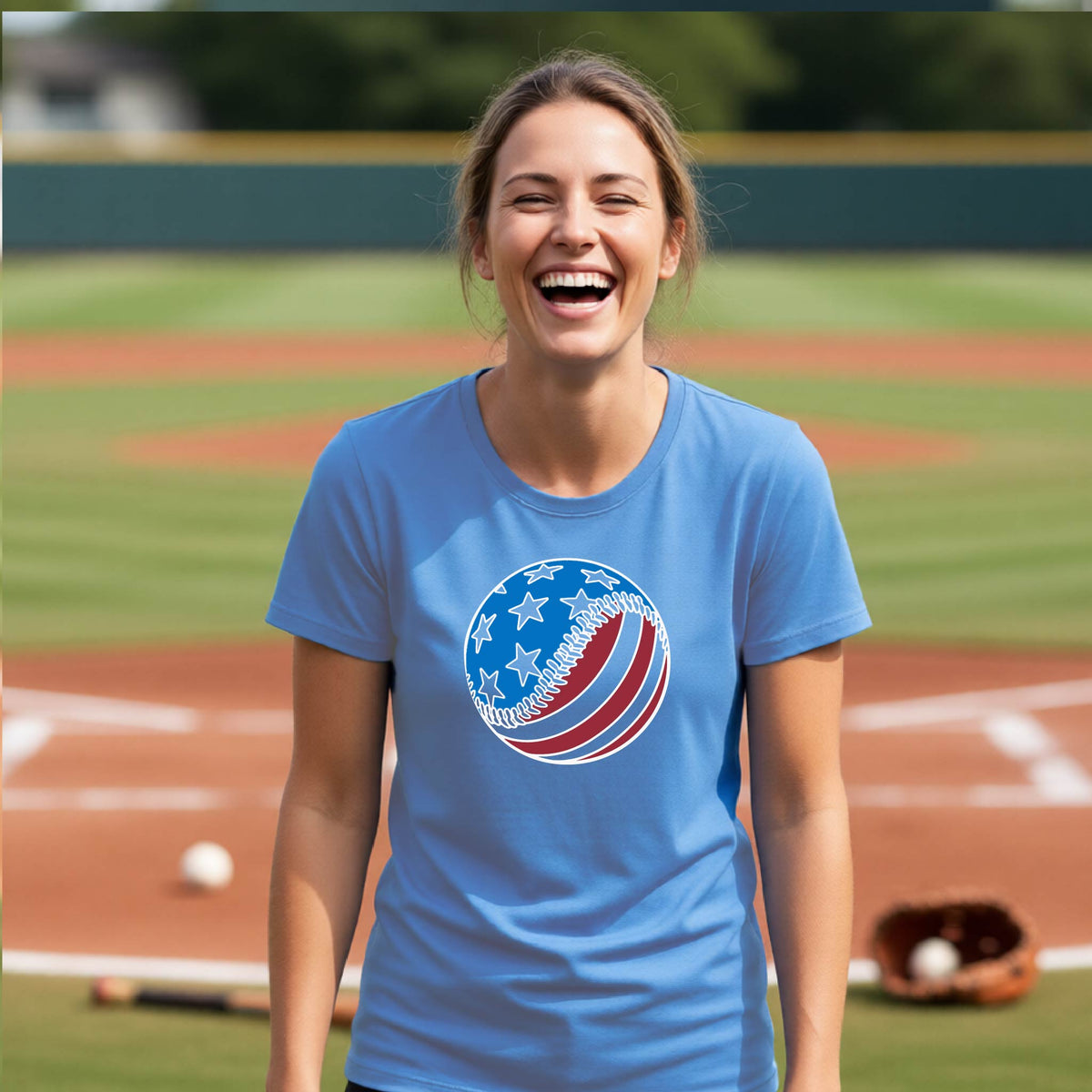 A woman smiling in front of a baseball diamond wearing a black t-shirt with a colored graphic of an American flag patriotic baseball with stars and stripes printed on the front.