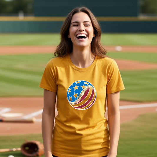 A woman smiling in front of a baseball diamond wearing a gold t-shirt with a colored graphic of an American flag patriotic baseball with stars and stripes printed on the front.