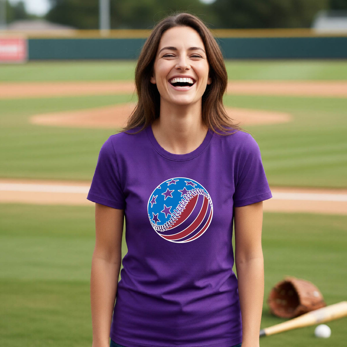 A woman smiling in front of a baseball diamond wearing a purple t-shirt with a colored graphic of an American flag patriotic baseball with stars and stripes printed on the front.
