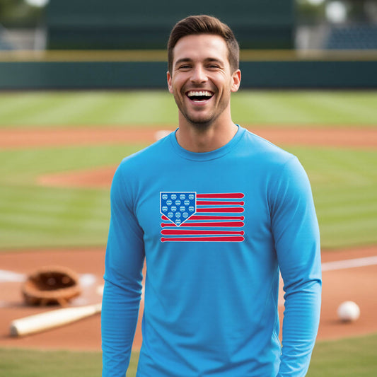 A man smiling in front of a baseball diamond wearing a bright blue long-sleeve t-shirt with a colored graphic of an American flag using baseball bats and balls and a home plate, printed on the front.
