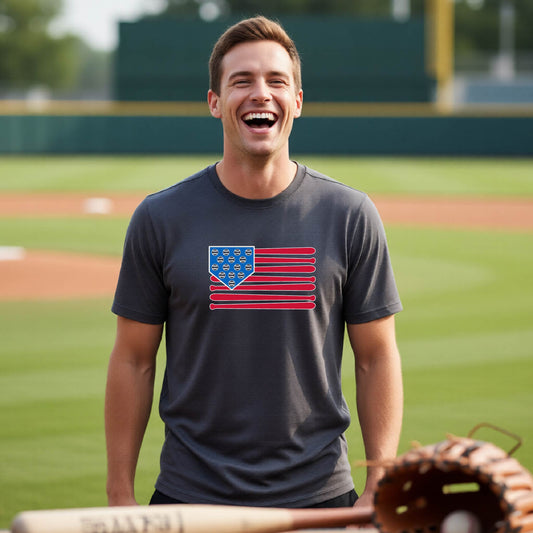 A man smiling in front of a baseball diamond wearing a dark gray short-sleeve t-shirt with a colored graphic of an American flag using baseball bats and balls and a home plate, printed on the front.