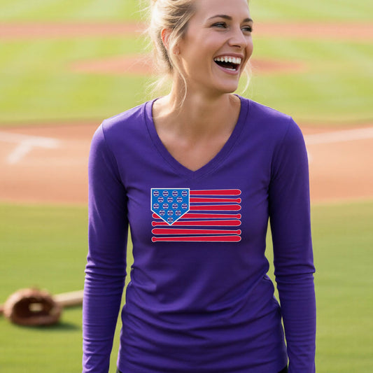 A woman smiling in front of a baseball diamond wearing a purple long-sleeve t-shirt with a colored graphic of an American flag using baseball bats and balls and a home plate, printed on the front.