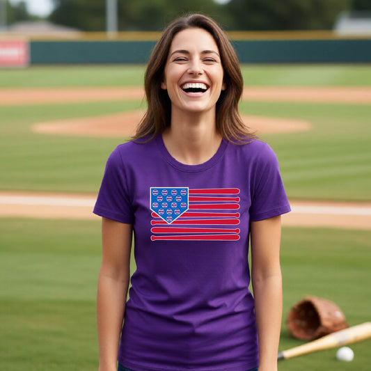 A woman smiling in front of a baseball diamond wearing a purple short-sleeve t-shirt with a colored graphic of an American flag using baseball bats and balls and a home plate, printed on the front.