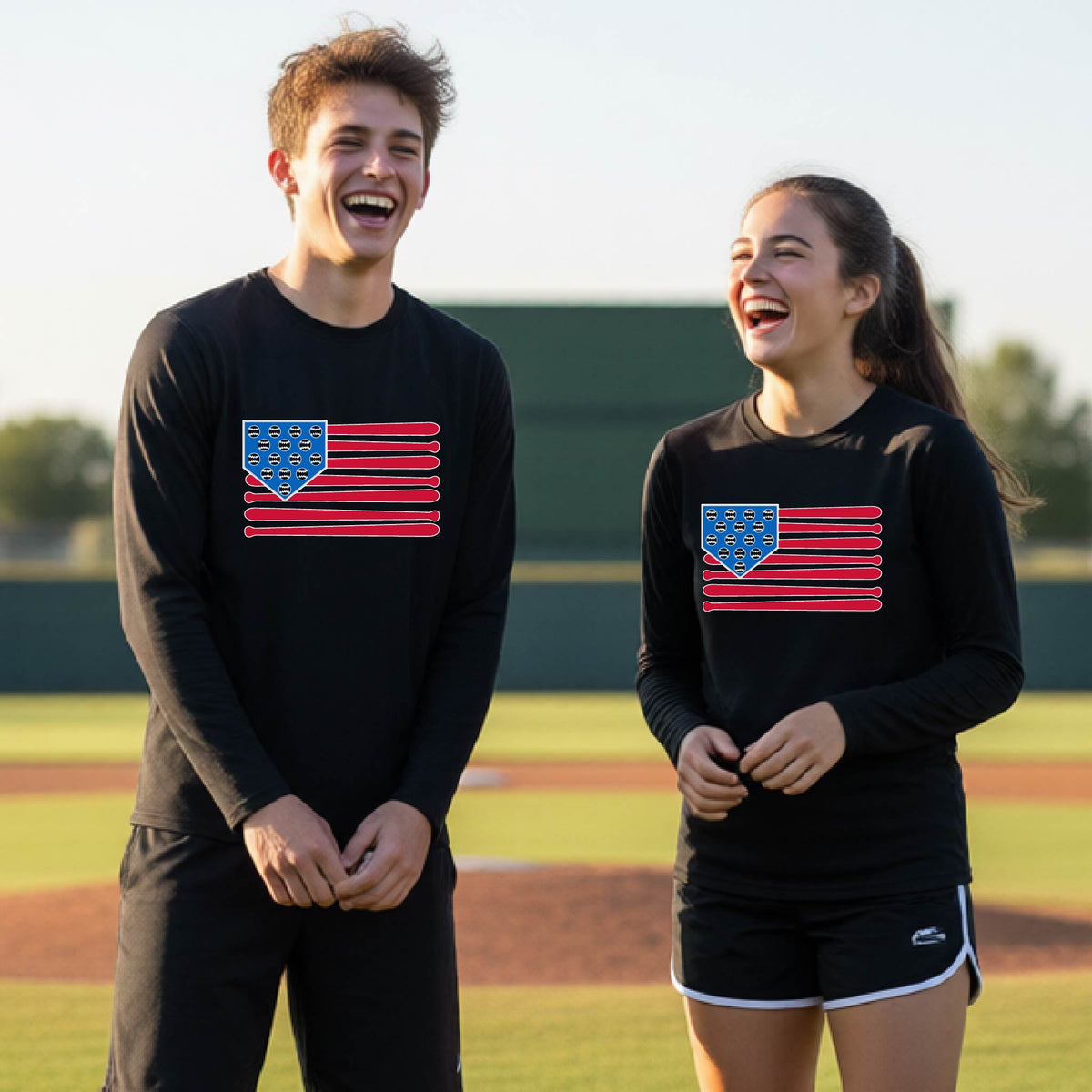 Two young athletes wearing matching shirts smiling in front of a baseball diamond wearing black long-sleeve t-shirts with a colored graphic of an American flag using baseball bats and balls and a home plate, printed on the front.