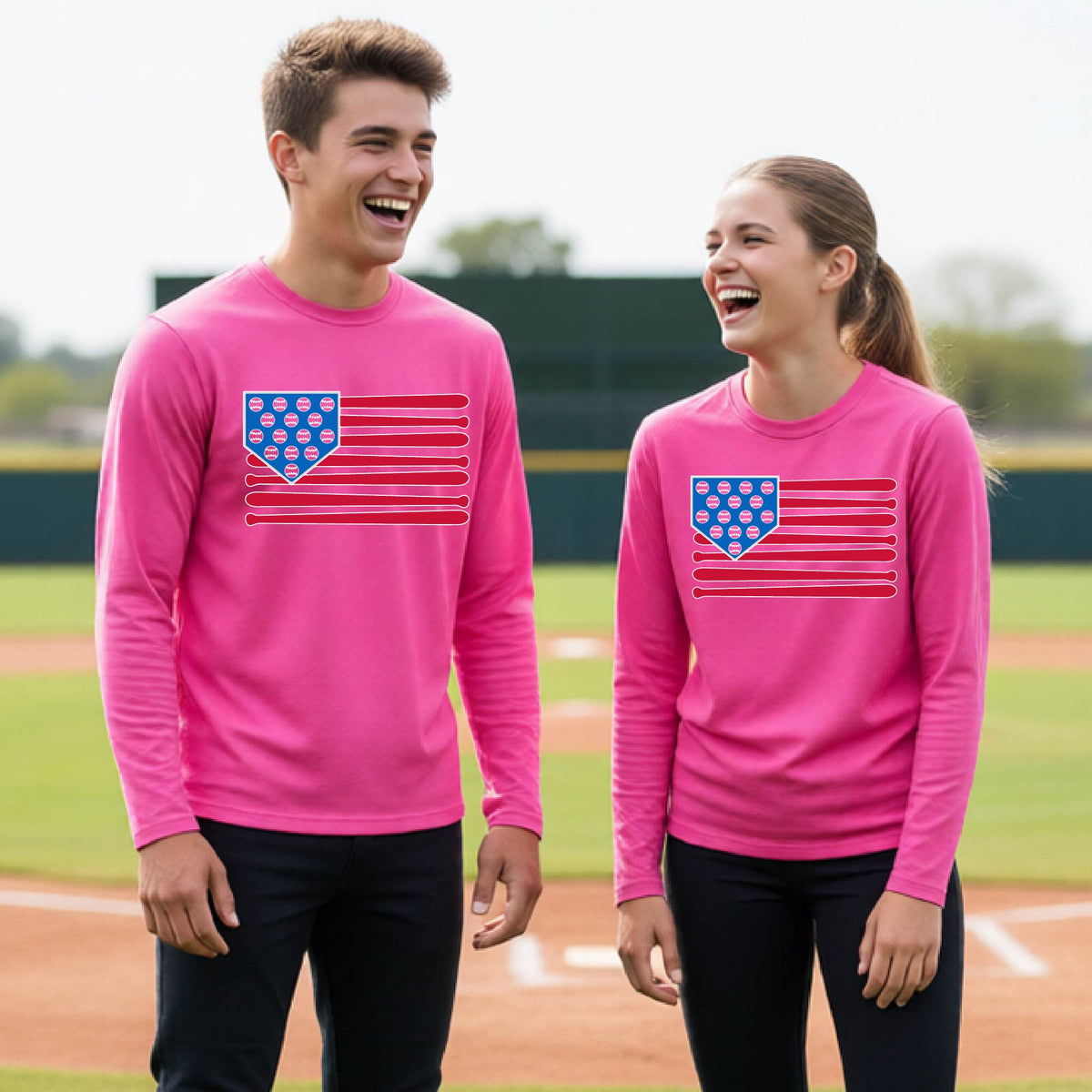 Two young athletes wearing matching shirts smiling in front of a baseball diamond wearing pink long-sleeve t-shirts with a colored graphic of an American flag using baseball bats and balls and a home plate, printed on the front.
