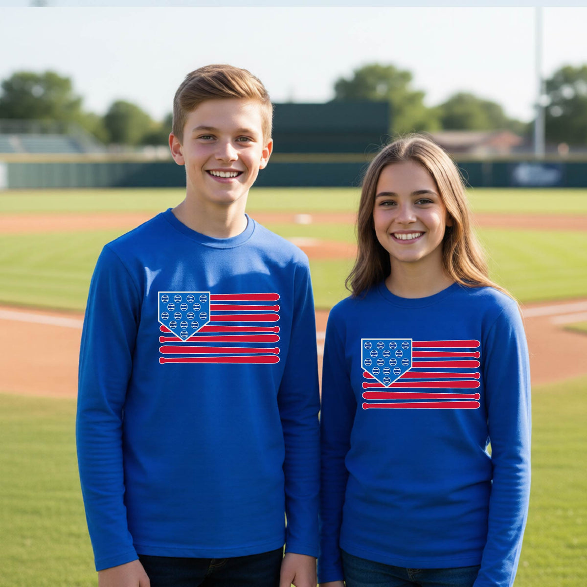 Two young athletes wearing matching shirts smiling in front of a baseball diamond wearing royal blue long-sleeve t-shirts with a colored graphic of an American flag using baseball bats and balls and a home plate, printed on the front.
